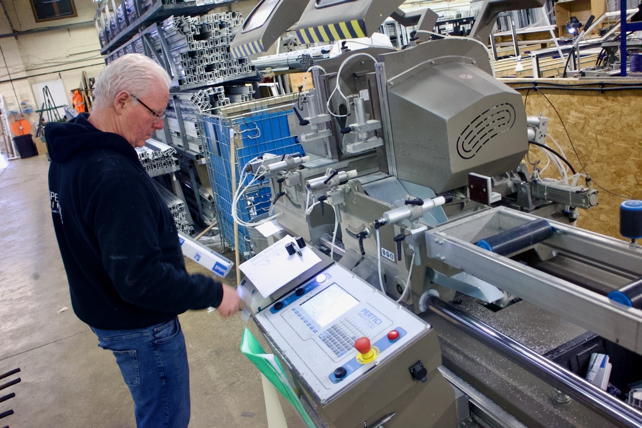 An older man with white hair and glasses works at a machine in a manufacturing facility. The machine appears to be a precision cutting or assembly device. The background is filled with shelves of metal profiles and other materials.