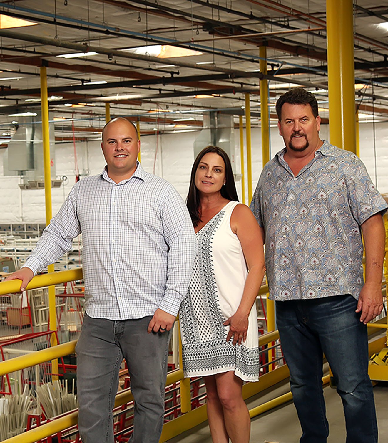 Three Avanti employees standing on a yellow-railed balcony overlooking a large manufacturing floor. The factory is spacious with various equipment and structures visible in the background.