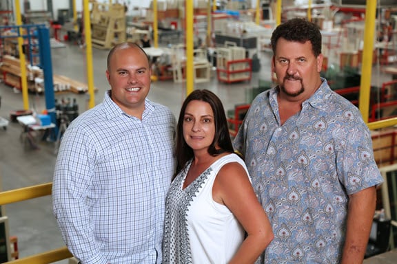 Close-up of three Avanti employees standing on a yellow-railed balcony inside a factory. The manufacturing floor and equipment are visible in the background.