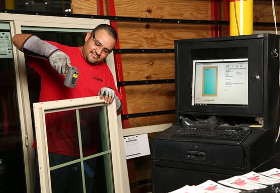 An Avanti worker in a red shirt and protective gloves assembling a window frame. The worker is using a power tool while a computer monitor next to him displays the window's design specifications.