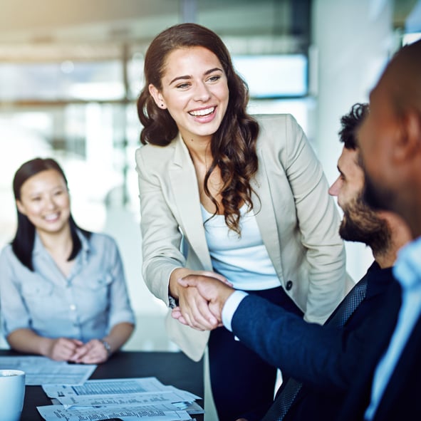 A professional woman with long brown hair, wearing a beige blazer and white blouse, is standing and smiling while shaking hands with a client across a conference table. Another woman, seated and smiling, is watching the interaction. The setting is a modern office with large windows and paperwork spread out on the table, signifying a successful business meeting and partnership.