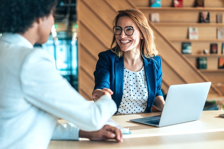 A customer success manager with glasses and a blue blazer is smiling and shaking hands with a client, symbolizing a successful agreement or partnership. She is sitting at a table with a laptop in front of her, in a modern office setting with wooden shelves in the background.