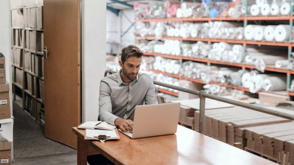 A man sitting at a table in a warehouse using a laptop.