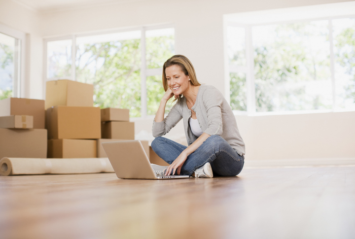 A woman sitting on the floor with a laptop, smiling as she works. The room is brightly lit with large windows, and there are moving boxes and a rolled-up carpet around her.