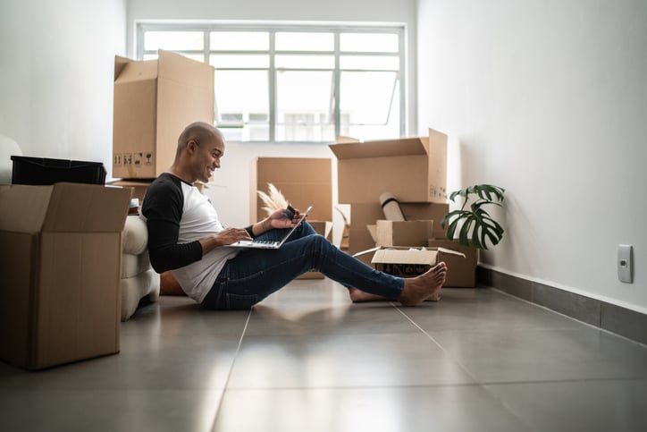 A man sitting on the floor with his back against a couch, working on a laptop. The room is filled with moving boxes, and a small plant is placed nearby.