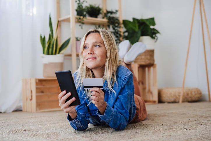 A woman lying on her stomach on the floor, holding a credit card and using a smartphone. The room is decorated with plants and wooden furniture.