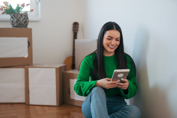 A woman sitting on the floor with her back against a wall, smiling while using a tablet. The room has several moving boxes and a small plant in the background.