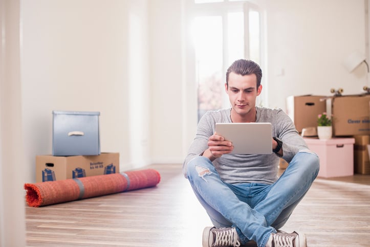 A man sitting cross-legged on the floor, using a tablet. The room is filled with moving boxes, a rolled-up carpet, and bright natural light from a window.