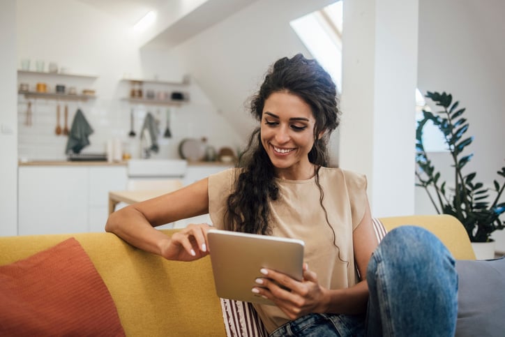 A woman sitting comfortably on a yellow sofa, using a tablet in a bright, modern home environment.