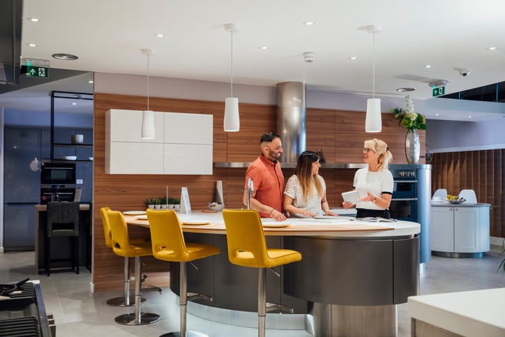 A couple consults with a kitchen designer in a showroom. They are standing around a large island with yellow bar stools, discussing design options with the designer, who is holding a tablet.