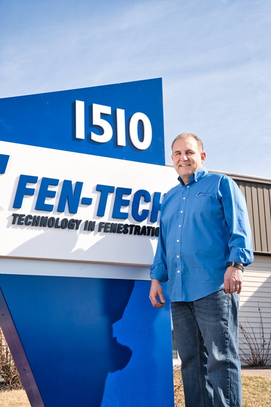 A man wearing a blue shirt stands in front of Fen-Tech sign