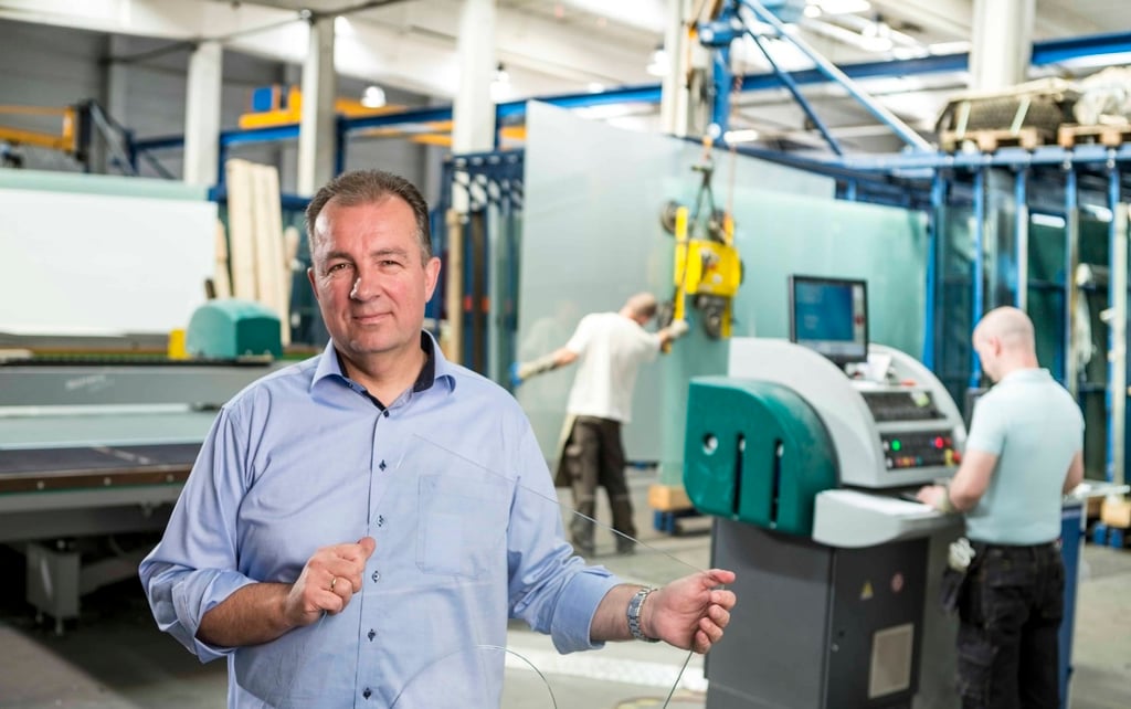 A man stands in front of a factory machine holding a sheet of glass