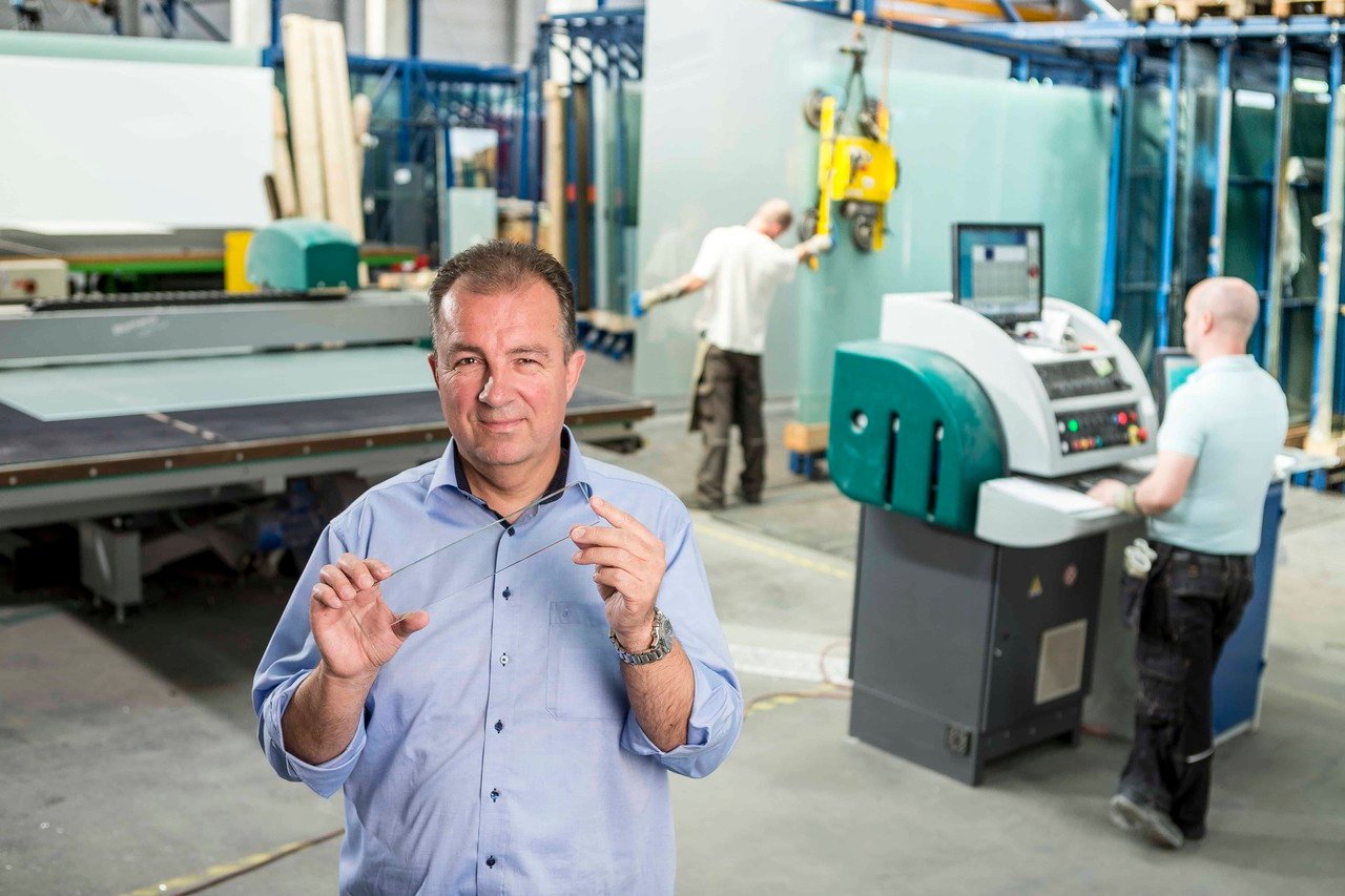 A man stands in front of a factory machine holding a piece of glass