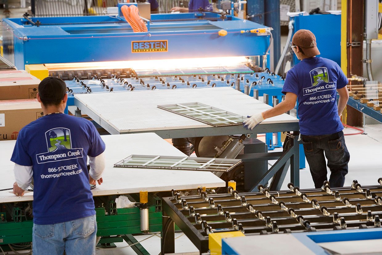 Two Thompson Creek employees wearing branded blue T-shirts work in a production line, placing window components on conveyor belts under industrial lighting. The background shows machinery labeled "BESTEN" for manufacturing processes.